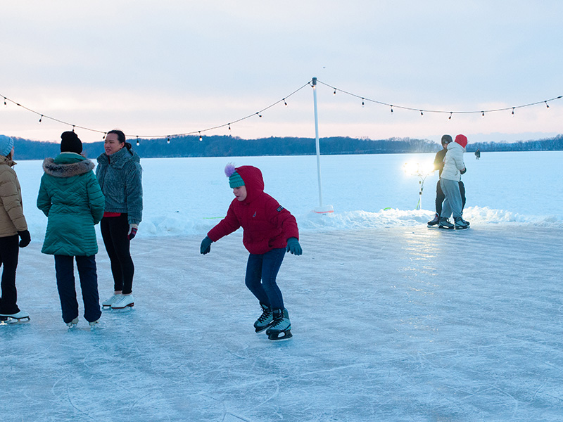 Ice skaters skate on frozen Lake Mendota with decorative lights in the background.